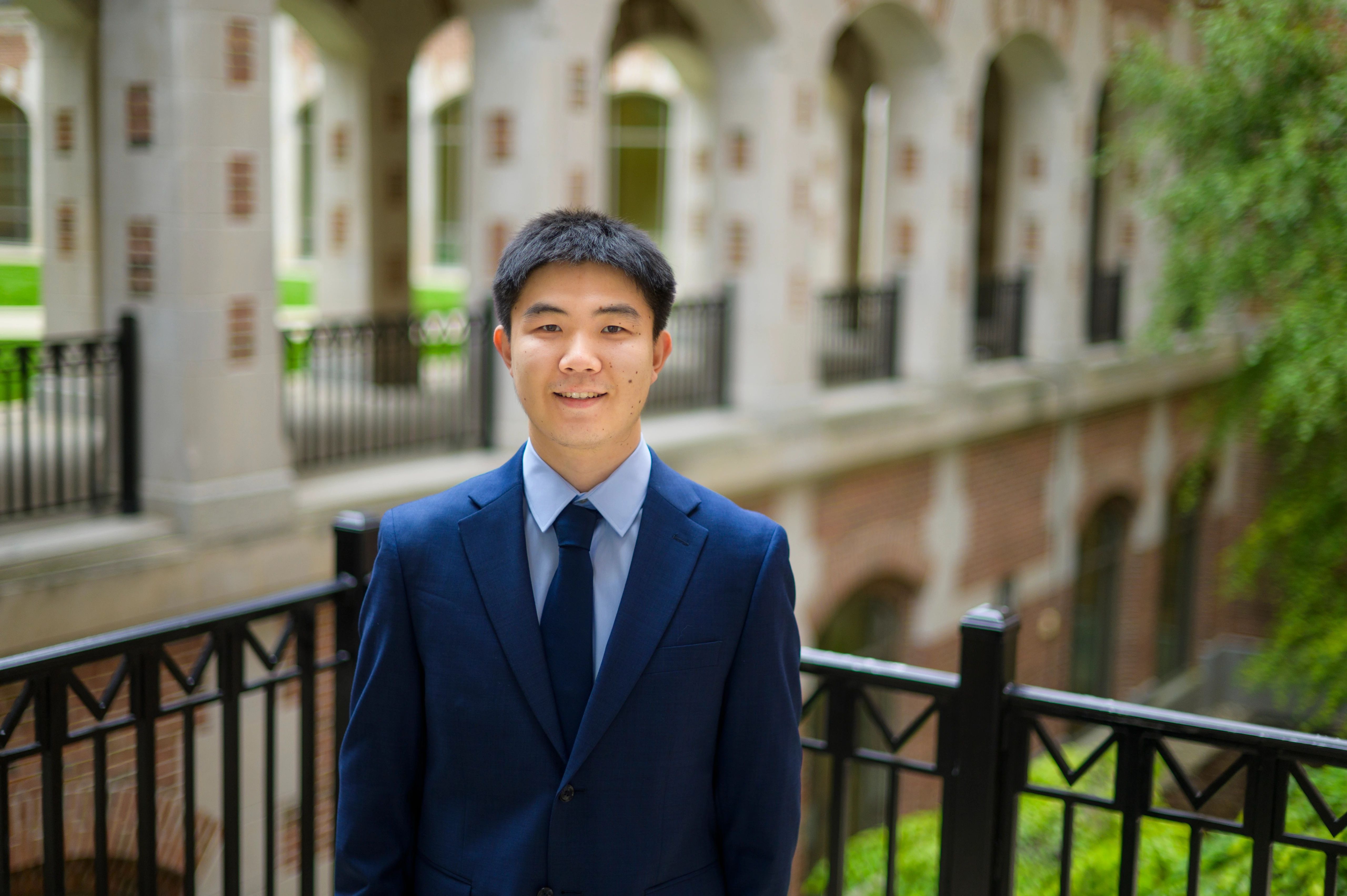 Jiaxin Pei wearing a blue suit and tie, standing outside and smiling at the camera.