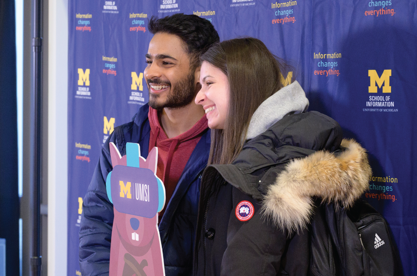 Two people standing together posing and smiling in front of a UMSI branded backdrop. They are holding a cutout of a purple squirrel holding an acorn and wearing a VR helmet. The helmet has the UMSI logo on it.