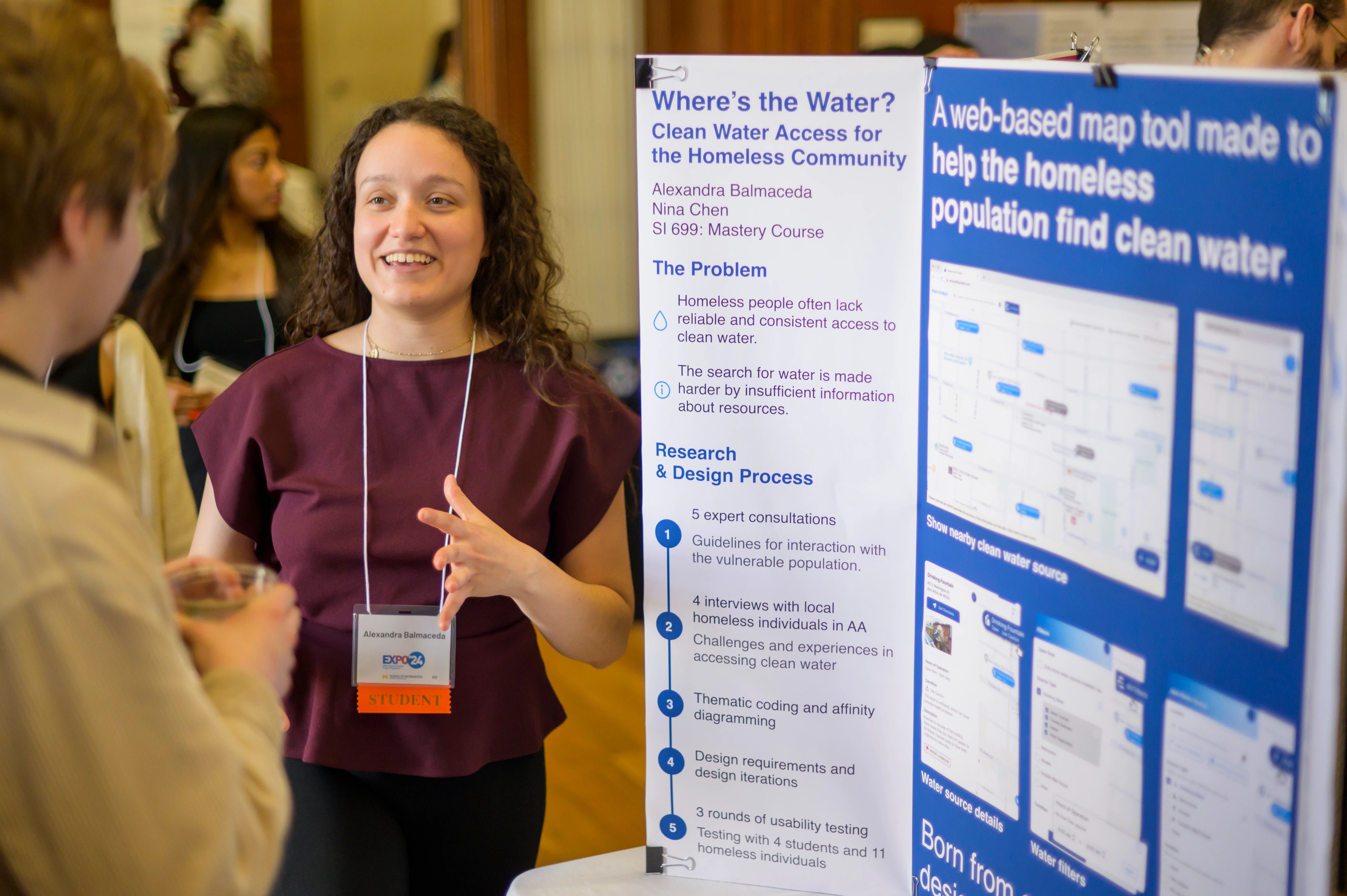 A student speaks with an Expo guest in front of a trifold poster about expanding clean water access for the homeless community.