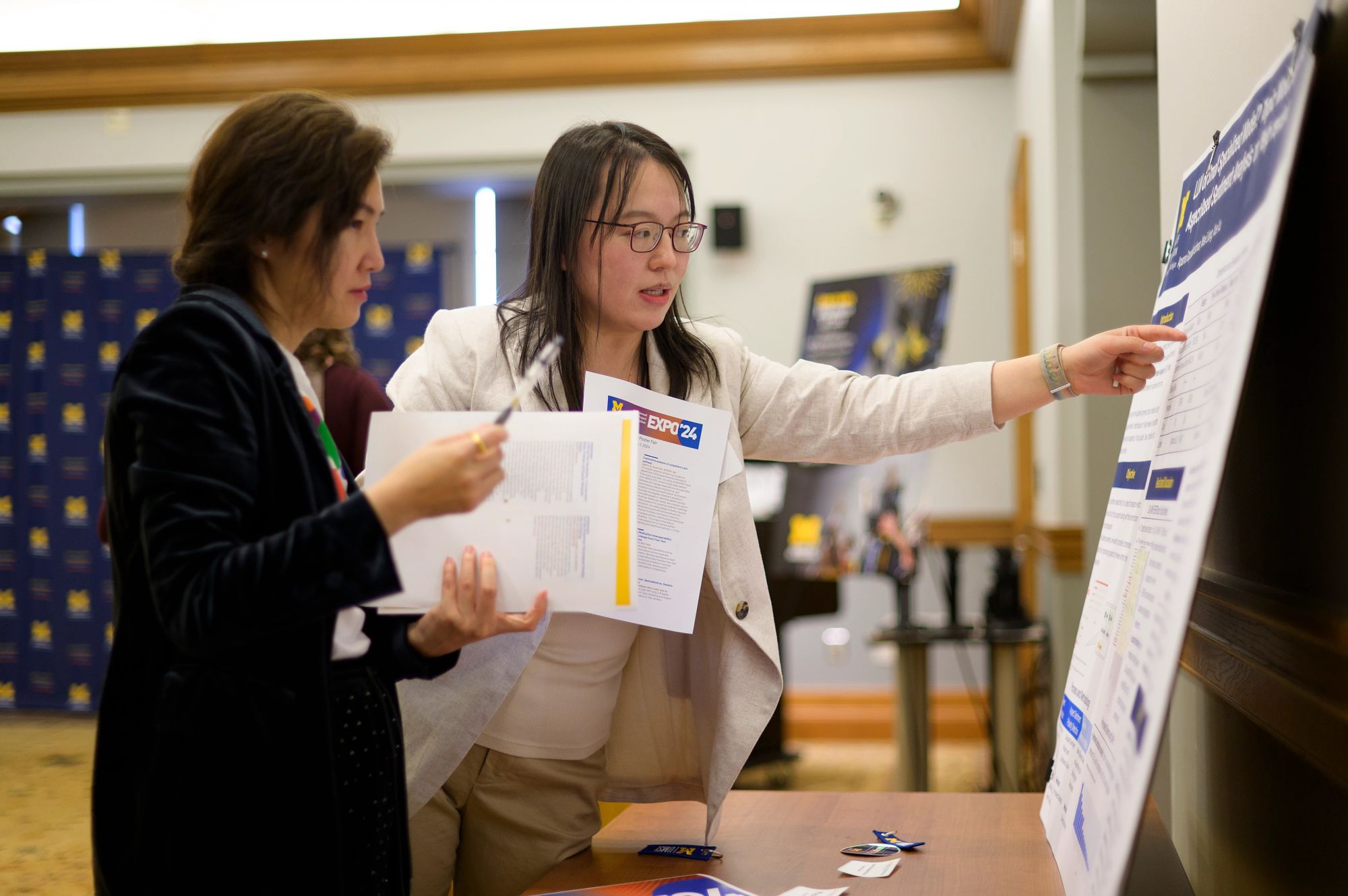 A student points to a poster while another student stands beside her, holding a project brochure.