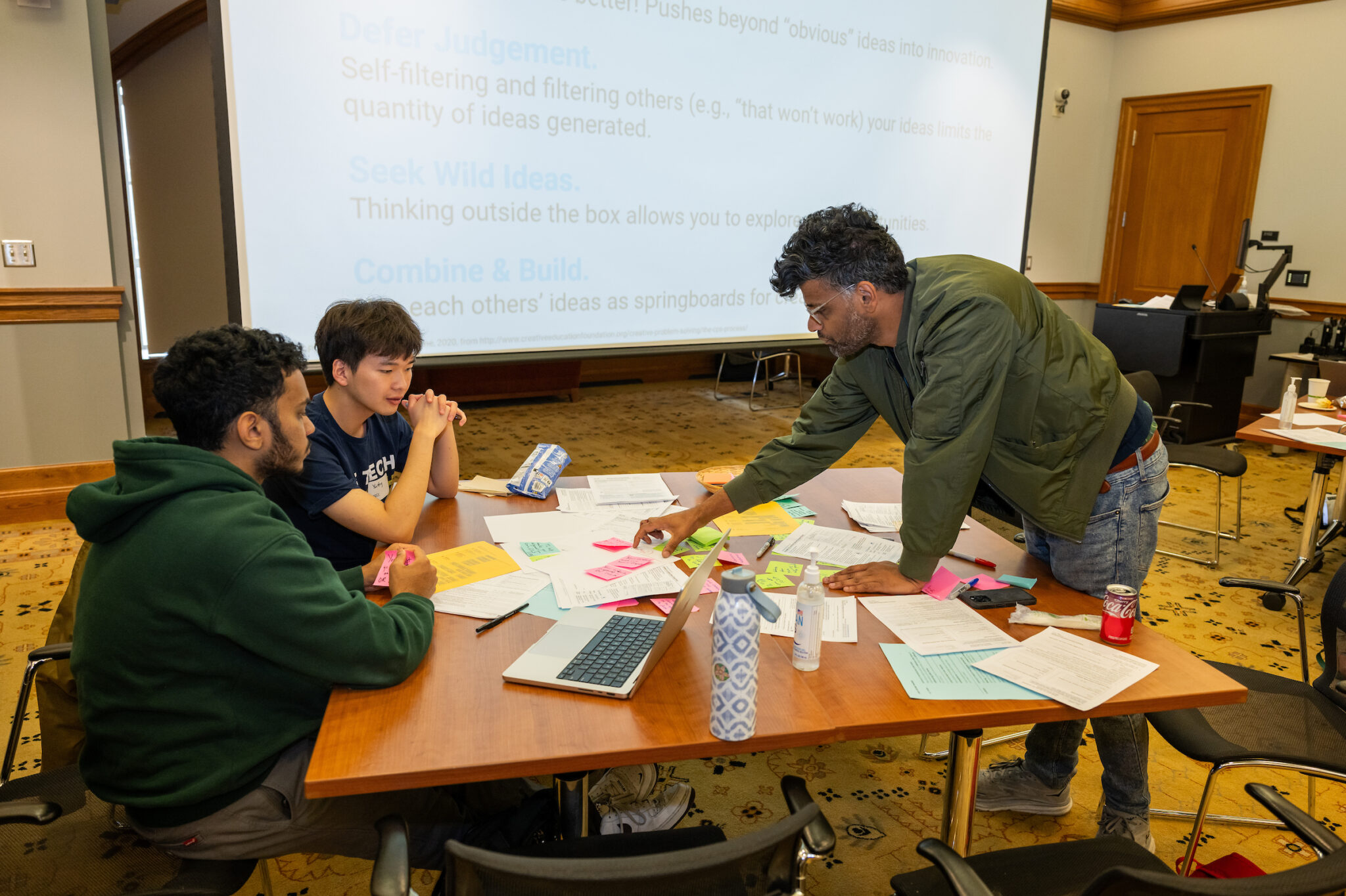 A workshop participant stands over a table pointing to a form while two workshop participants look on, seated