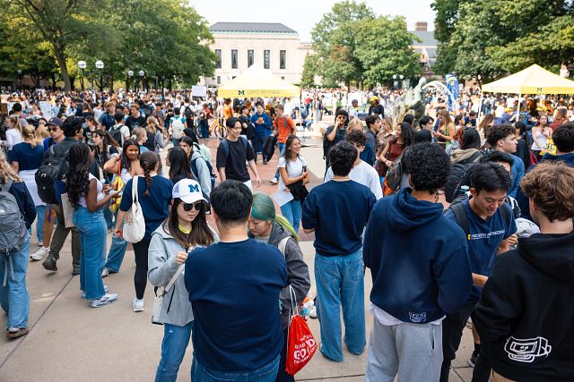 A large crowd gathered outside. There are tents and an academic building in the background.
