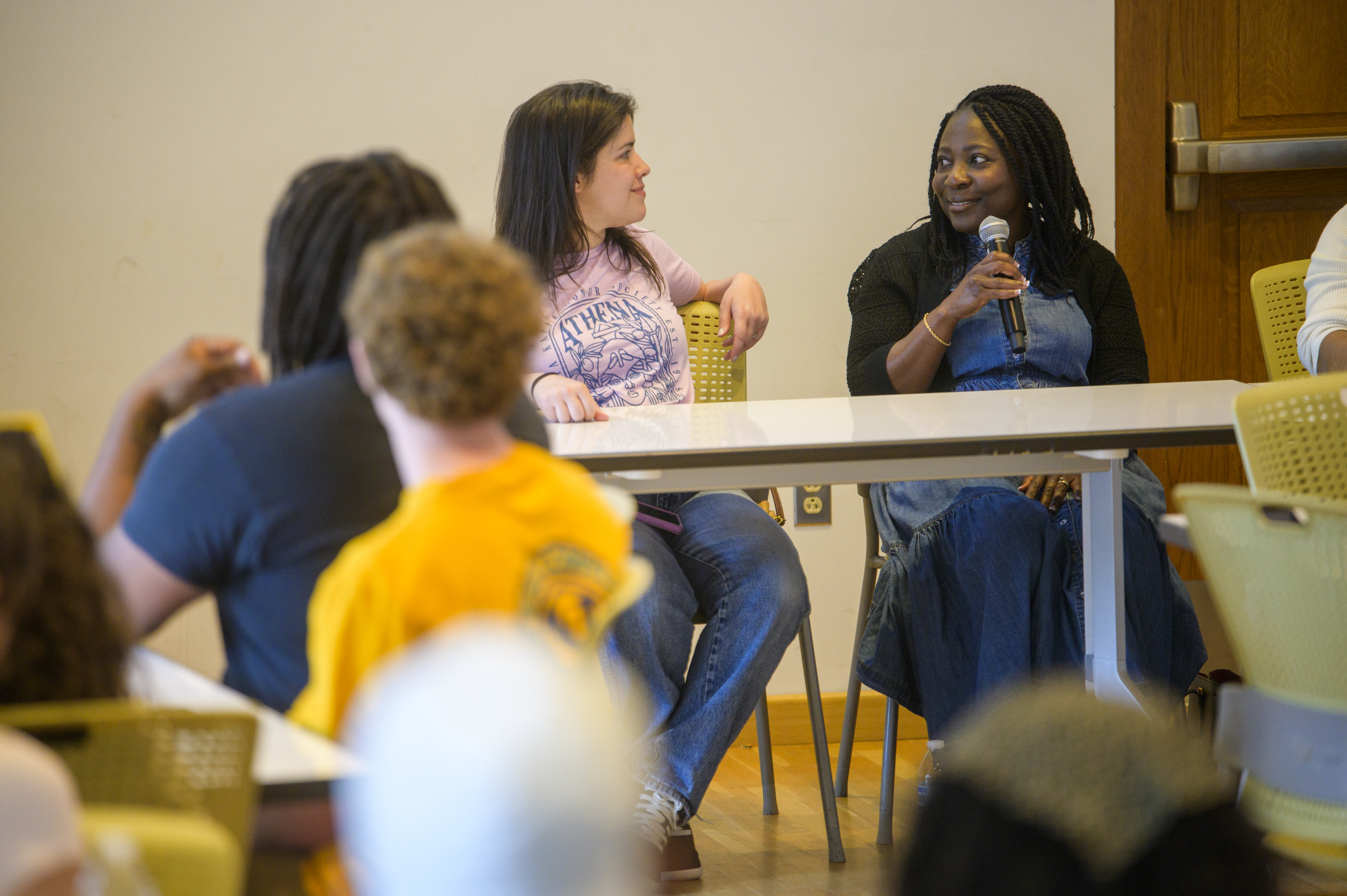 Two students sit at a long table in front of a crowd of onlookers, with one holding a microphone as the other looks on