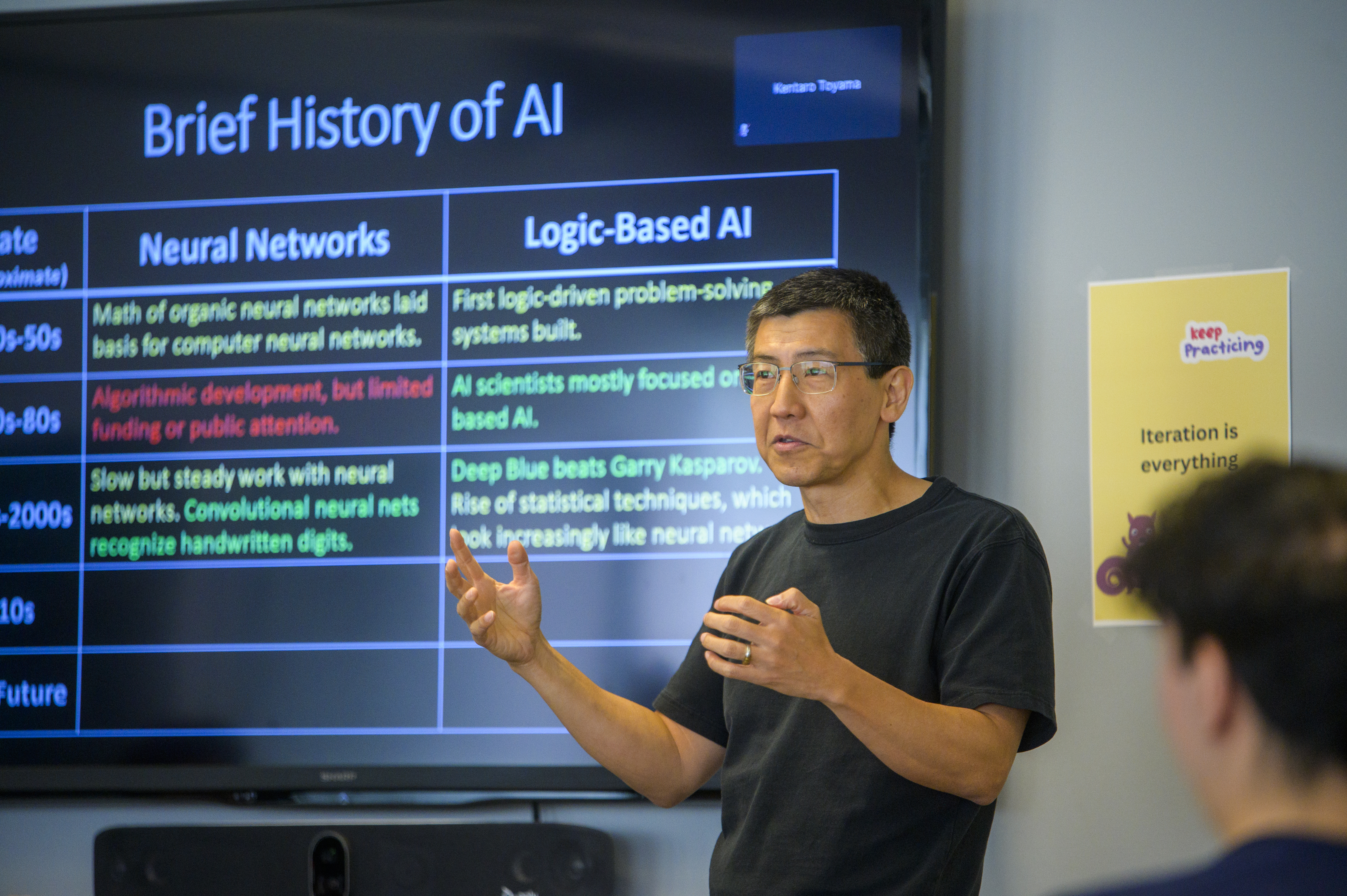 Professor Kentaro Toyama gestures with his hands while standing in front of a screen titled Brief History of AI 