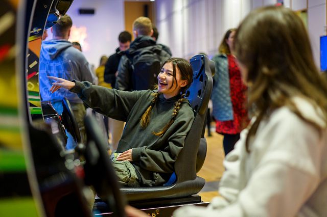 Two students sit at arcade cabinets with steering wheels attached to them.