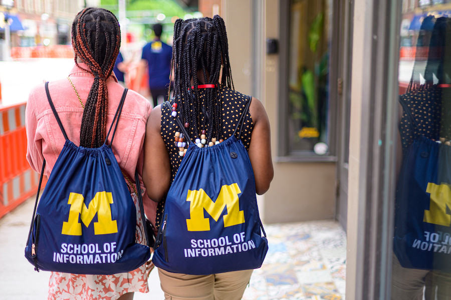 Two high school students walk beside each other, arms linked, on a sidewalk in downtown Ann Arbor. We see their backs; both wear backpacks that say School of Information. 