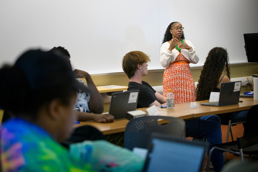 Four students are pictured in a classroom, heads turned to listen as Megan Threats, assistant professor, lectures in front of a whiteboard. 