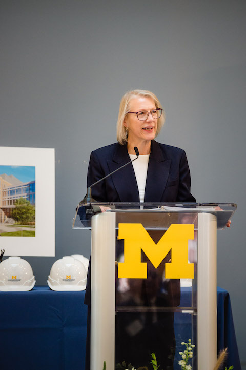 ​​​Interim dean Elizabeth Yakel, wearing a blue blazer, speaks from a podium at the groundbreaking ceremony for the new Leinweber Building. 