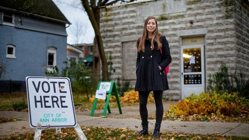 Shaelyn Albrecht standing in front of a polling location, there is a sign that says "vote here" to her right. 