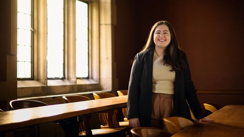 A photo of Julia Kassab posing behind a long wooden desk in an empty Law School classroom