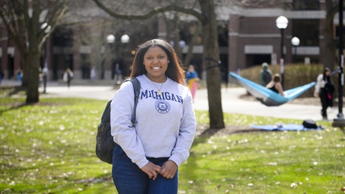A photo of Ambar Amoros-Gomez standing on the Diag wearing a U-M sweatshirt, with a hammock visible in the background