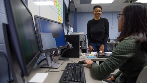 A student sitting at a desktop computer with two monitors talking to another student who is standing.