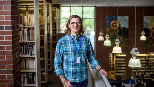 A person standing on a balcony in a public library looking over the first floor. There are bookshelves and a window behind him.