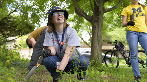 A student kneeling in the grass and holding a gardening spade.