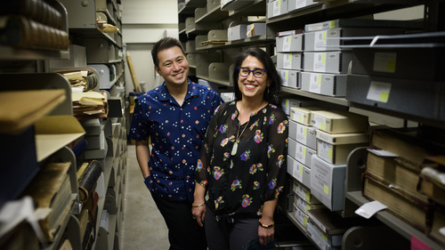 Two people smiling and posing between shelves of boxes in an archive.