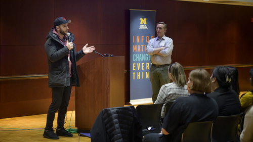 A person giving a talking next to a podium. There is a UMSI banner next to him.
