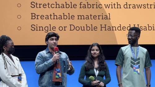 Four students stand on a stage in front of a digital slide. One speaks thoughtfully into a microphone 