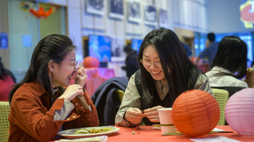 Two students laugh together at a table with a red tablecloth and paper lanterns on it.