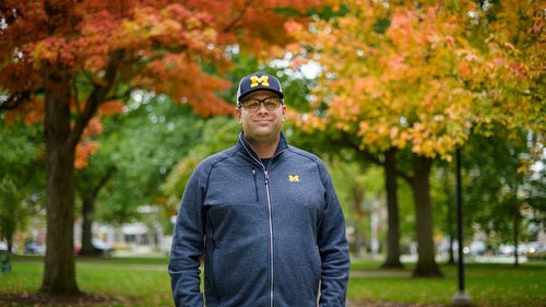 Kyle Bylin stands on the U-M campus with fall foliage in the background.
