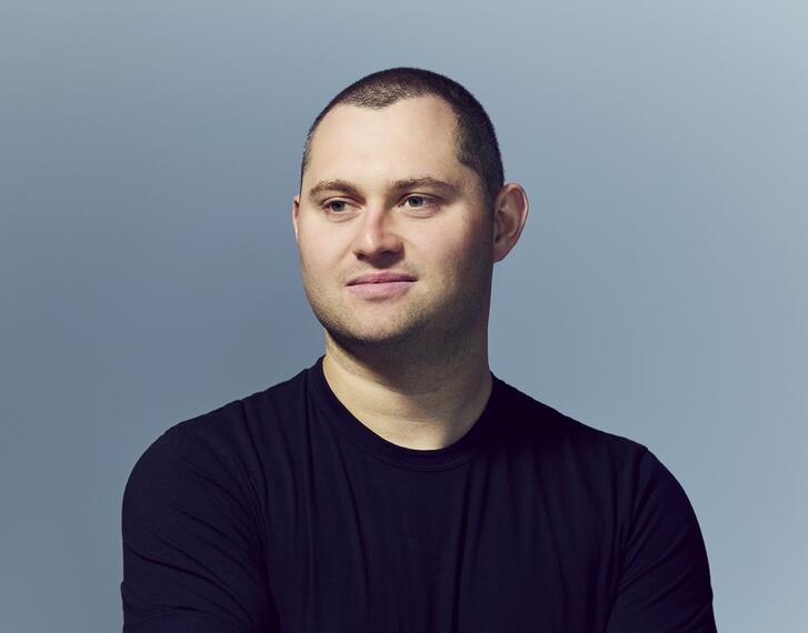 A headshot of Mark Gurman posing with arms crossed against a slate gray background