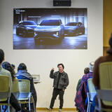 A person giving a presentation to a room full of people. A photo of three cars is projected on a screen above him.