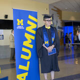 A student wearing graduation regalia standing next to a banner that says "Alumni"