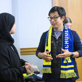 Two people talking together. One of them is wearing a University of Michigan scarf.