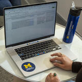 Hands on a laptop with University of Michigan School of Information stickers. The stickers read “mad science mode,” “change everything,” and “plays well with data.”