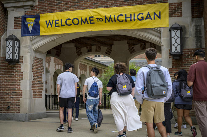 About 10 students walk into an outdoor brick passageway that is decorated with a maize and blue banner that reads "Welcome to Michigan." Two of the students wear UMSI-branded backpacks.