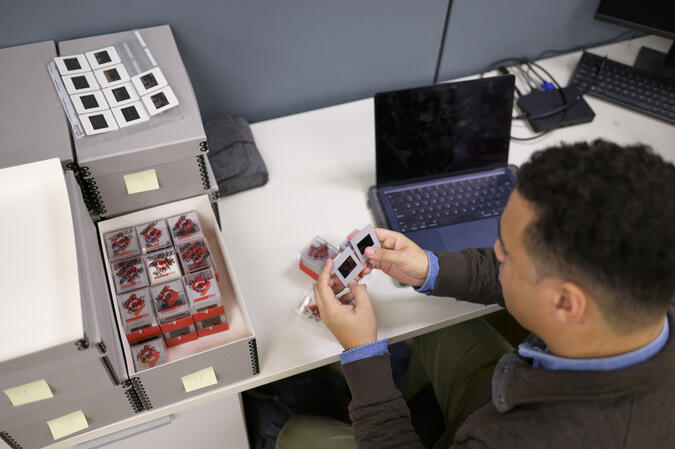 A student sitting at a desk with a laptop and sorting through a box of film slides.