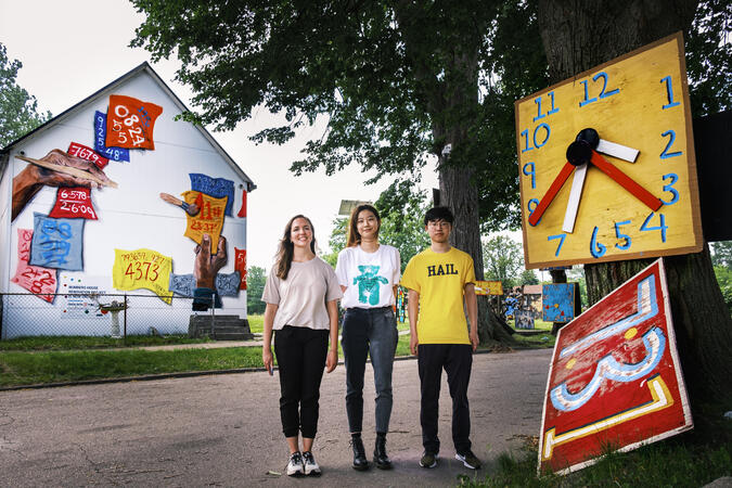 Three students stand in front of a house with a mural painted on the side and next ot a tree with a handmade clock hanging on the front.