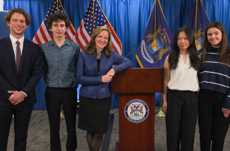 Five people stand in a line next to a podium. There are American flags and State of Michigan flags in the background.