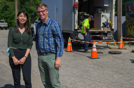 Two people posing and smiling in front of a group of construction workers and a truck.