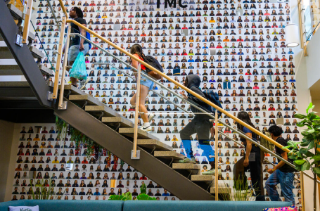 Students climbing a stairway in front of a wall covered in hundreds of small headshots.