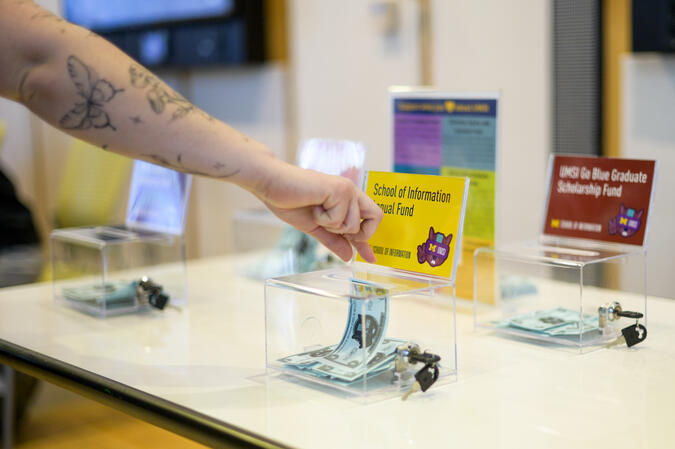 A hand dropping a slip of paper into a clear box with a label that says "School of Information Annual Fund." There are three other boxes on the table.