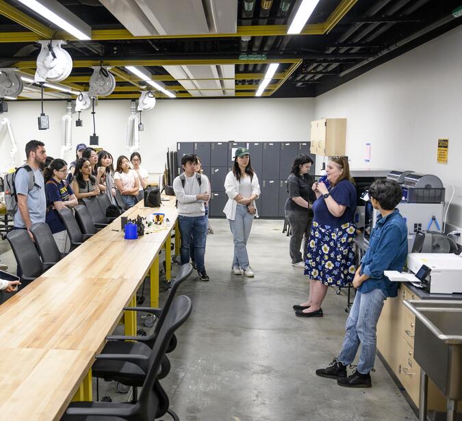 Students standing around long wooden tables listening to another person talk. They are standing in front of a counter covered in small machines.