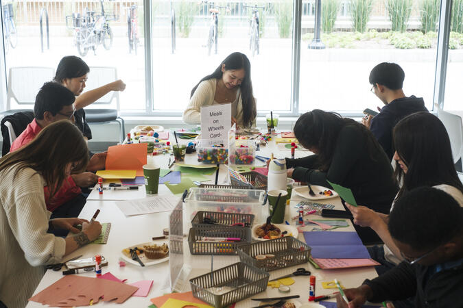 Students working at a table with construction paper and craft supplies. There is a large window behind them.