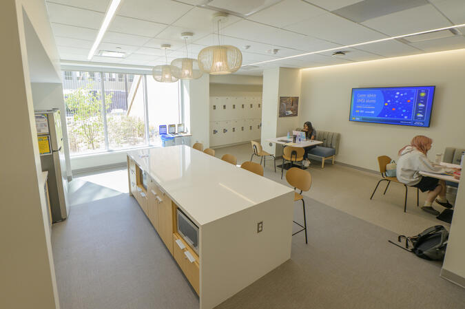 A brightly lit room with a kitchen island in the middle, a wall of windows, a set of lockers, and two tables with students sitting and working.