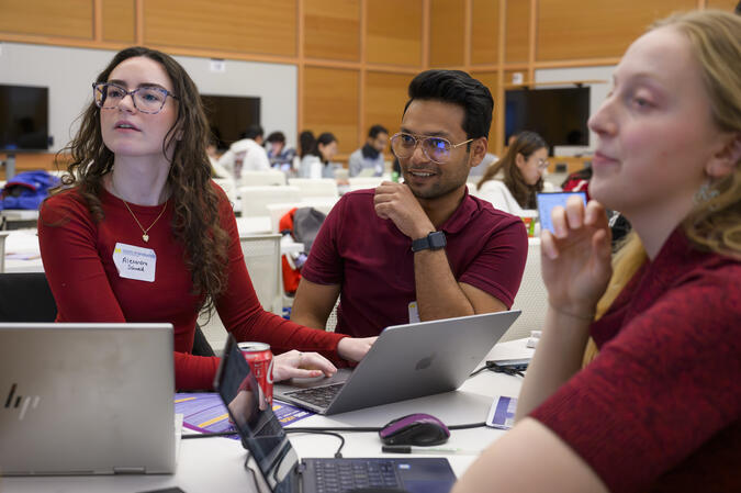 Three teammates, all wearing red, smile and glance to the left. Each has a laptop on a table in front of them in a large academic room