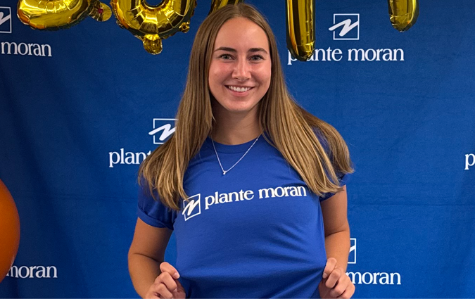 A student wearing a shirt with the Plante Moran logo standing in front of a background with the Plante Moran logo on it.