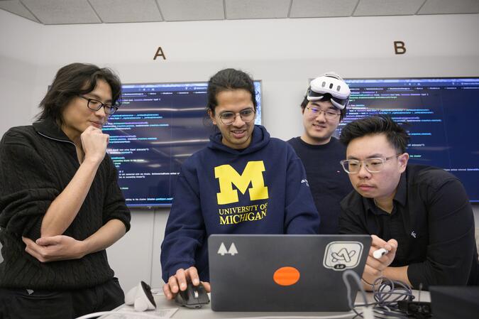 Four people standing around a laptop. One is wearing a VR headset pushed up on their forehead. There are two screens on the wall behind the people with code on them.