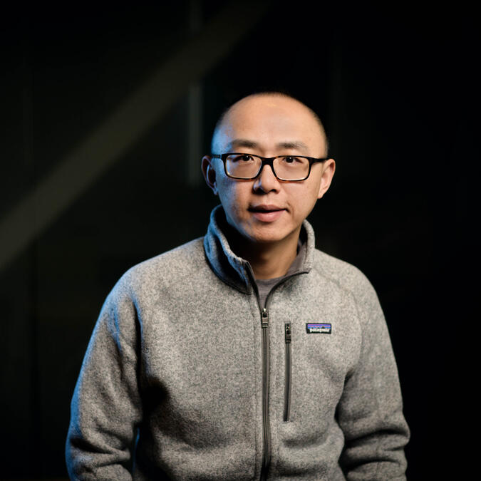 Jiayu Zhou wears a sweater and a black pair of glasses and looks at the camera. 