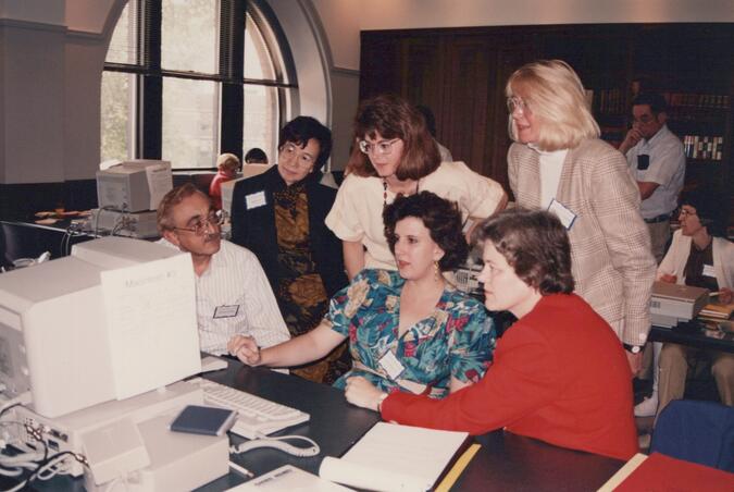 A film photograph of 6 people in retro clothes and hairstyles sitting and standing around an old desktop computer.