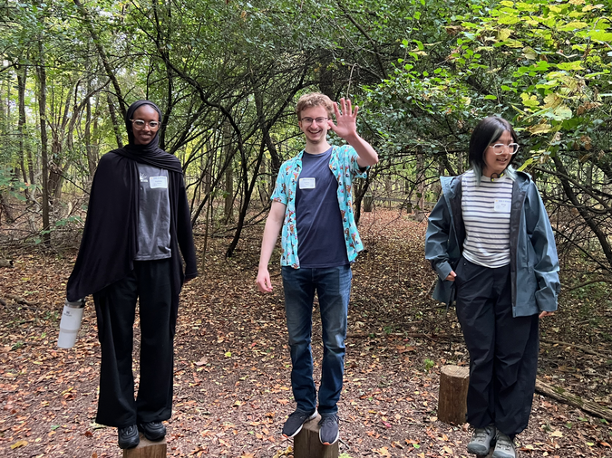 Three students outside, standing on top of logs.