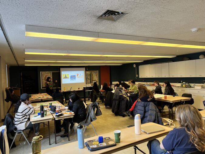 Students sitting in a classroom at long tables, watching a presenter at the front of the room.