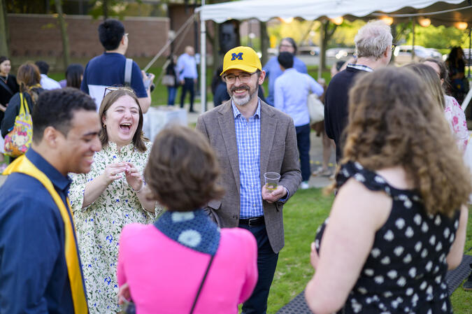 A group of people standing outside together and laughing. The person in the middle is wearing a U-M hat.