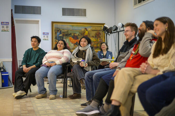 A small audience of students faces forward as if watching a performance, in a warmly lit carpeted room with art on the wall