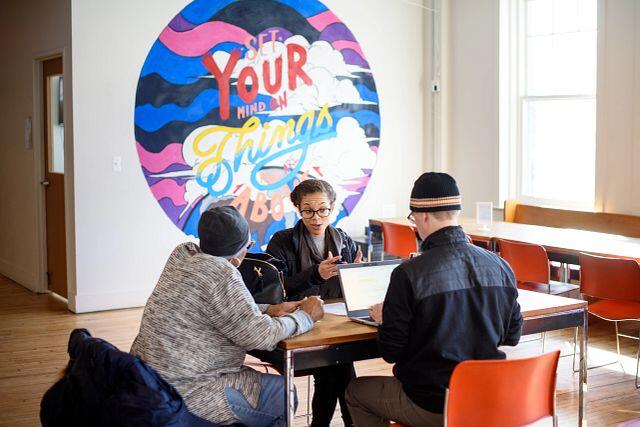 Three people sit at a table. One is on a laptop and another is midsentence.
