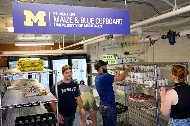Four people in a room with shelves of food and a refrigerator case. The sign above them says Maize & Blue Cupboard.