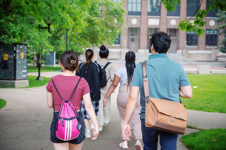 Five community college students are pictured walking ahead of the camera, surrounded by campus greenery and buildings, as they tour the U-M campus.